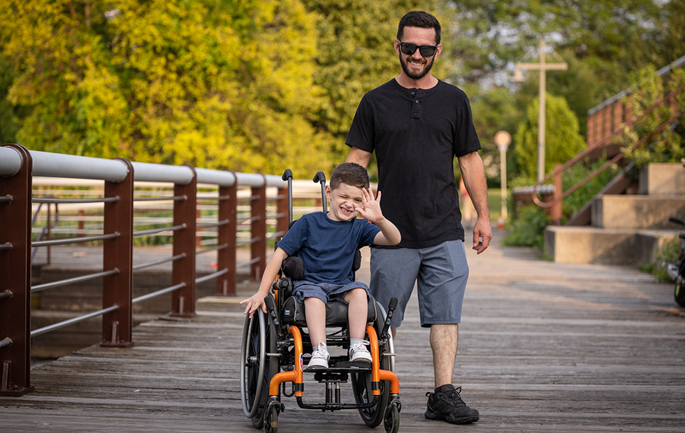 A father walks next to his son, waving and smiling in a wheelchair.